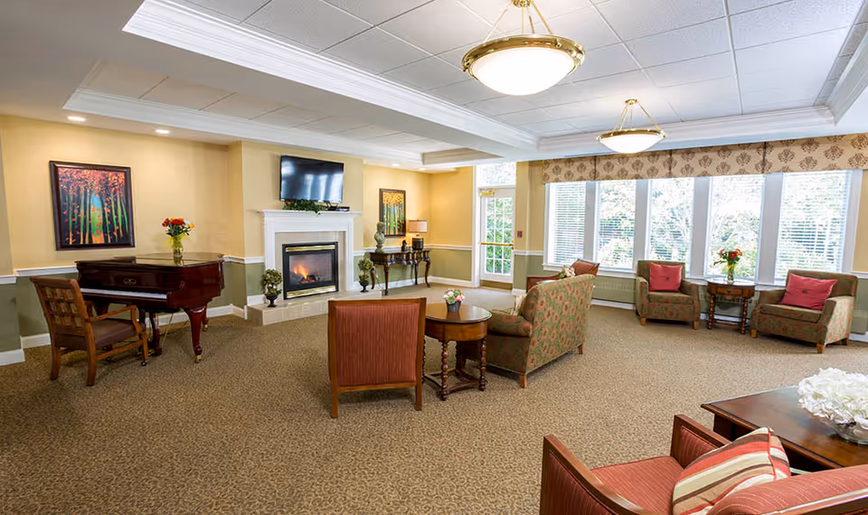 A spacious senior living common area with a fireplace and a wall-mounted TV above it. The room features multiple upholstered chairs and sofas arranged around small wooden tables with floral decorations. There is a grand piano with a chair on the left side and large windows on the right side letting in natural light. The walls are painted in warm tones with framed artwork and decorative lamps.