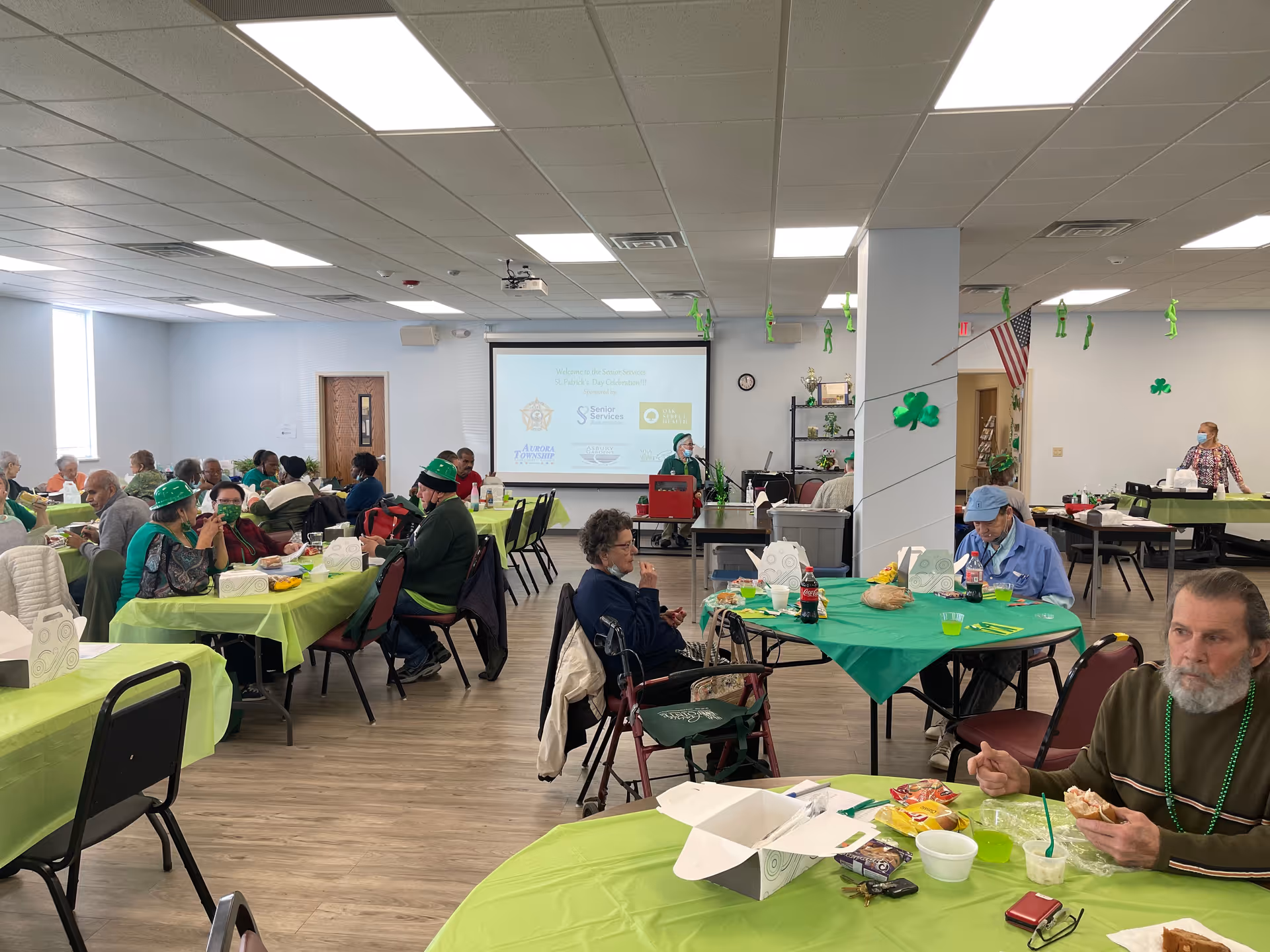 A group of elderly people seated at tables covered with green tablecloths in a large, well-lit room. The room is decorated with green shamrocks and other St. Patrick's Day themed decorations. Some people are wearing green hats and beads. There is a projector screen at the front of the room displaying logos and text, and an American flag is visible on the right side. The atmosphere appears to be a social gathering or event.