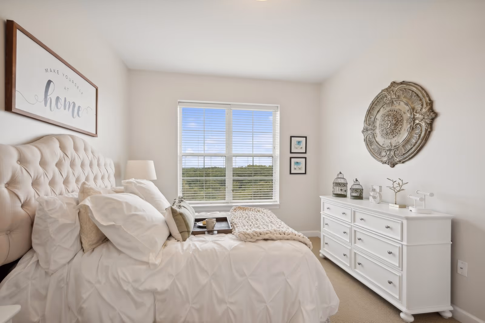 A bright and cozy bedroom with a large tufted headboard bed dressed in white bedding and multiple pillows. A tray with a cup and a book is placed on the bed. There is a white dresser with decorative items including birdcages and a small plant. A large ornate wall decoration hangs above the dresser, and two small framed pictures are on the wall next to a window with white blinds showing a green outdoor view. A framed sign above the bed reads 'Make Yourself at Home.'
