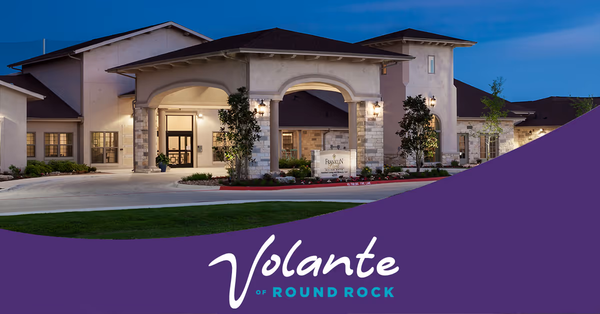 Front entrance of a senior living building lit at dusk with a purple banner across the bottom reading "Volante of Round Rock."