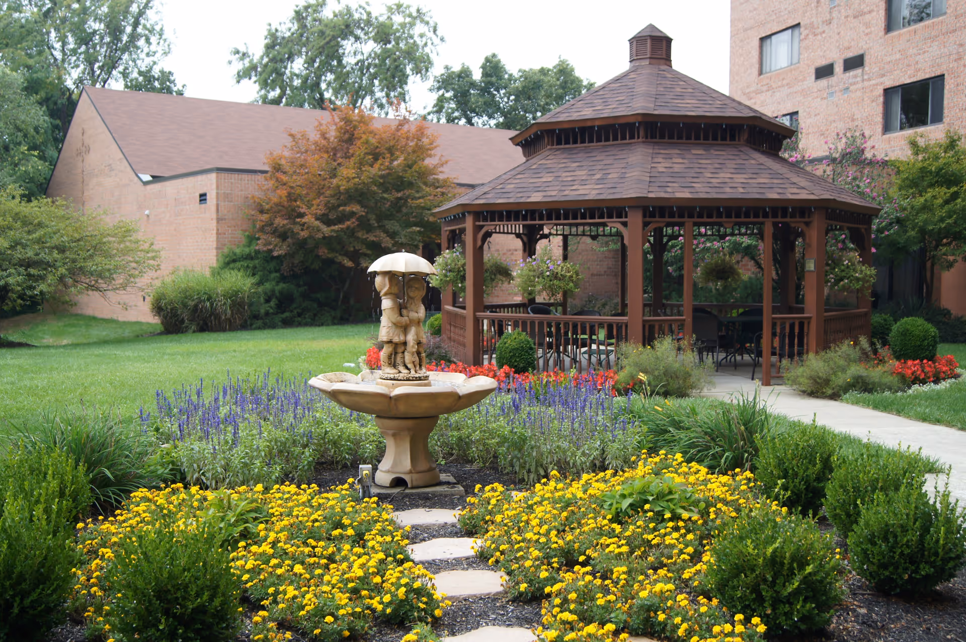 A garden area with a stone fountain featuring a sculpture of two children under an umbrella, surrounded by yellow and purple flowers and green shrubs. In the background, there is a brown wooden gazebo with hanging flower baskets and chairs inside, and brick buildings with windows.