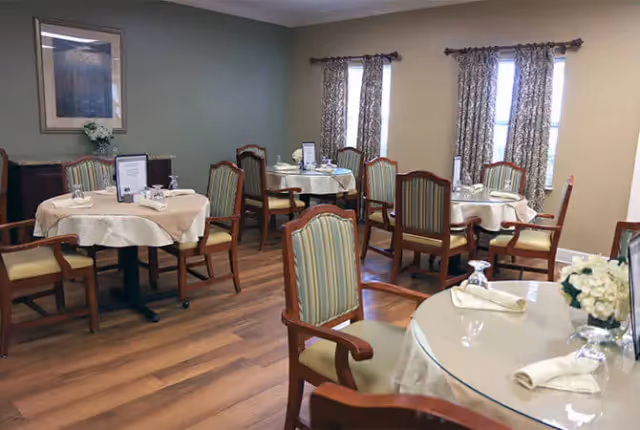 Dining room with several round tables covered with beige tablecloths, each set with napkins, glasses, and small flower arrangements. Wooden chairs with striped upholstery surround the tables. The room has wooden flooring, two windows with patterned curtains, and a framed picture on the wall.