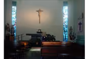 Interior view of a small chapel with wooden pews, an altar at the front, a crucifix mounted on the wall above the altar, stained glass windows on either side, and religious statues placed near the windows.