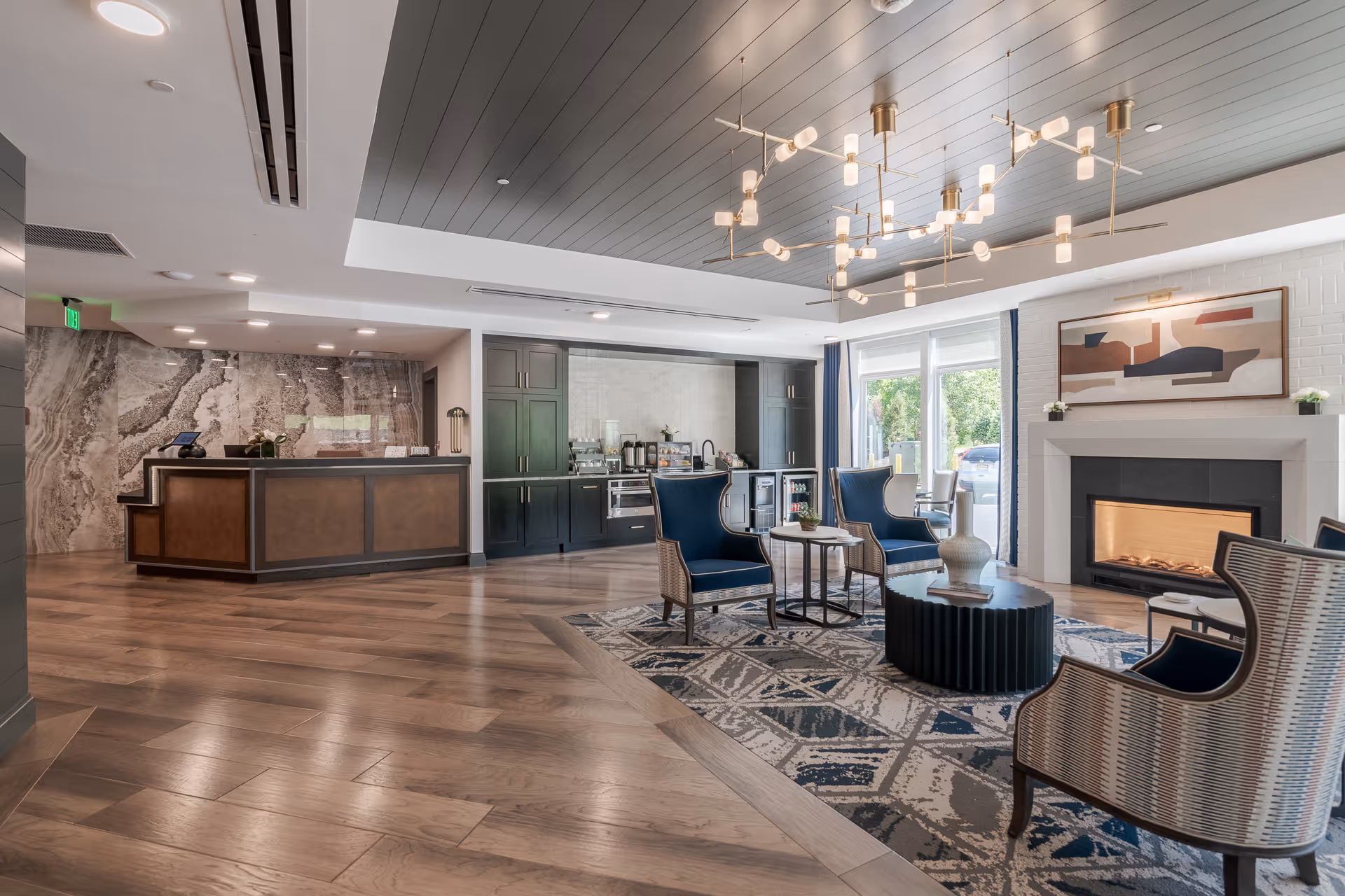 A modern and spacious senior living facility lobby with a reception desk on the left, a coffee and beverage station in the center back, and a seating area on the right featuring four armchairs around a coffee table on a patterned rug in front of a lit fireplace. Large windows allow natural light to brighten the room, and a contemporary chandelier hangs from the ceiling.