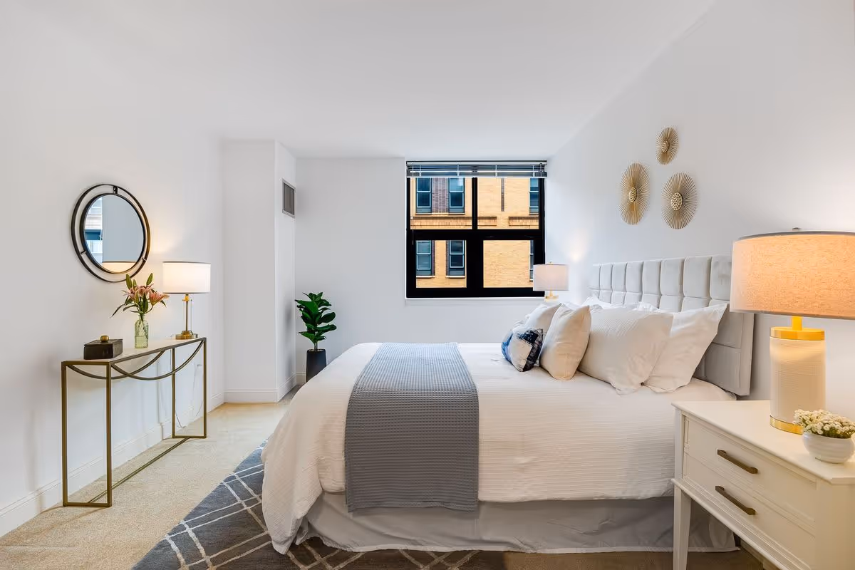 A bright and neatly arranged bedroom with a large bed featuring white bedding and multiple pillows. There is a gray throw blanket on the bed. On the right side, there is a white nightstand with a lamp and a small plant. On the left side, a decorative console table with a vase of flowers and a round mirror above it is placed against the wall. A window with a view of a brick building is centered on the back wall, and a potted plant is in the corner. The walls are white, and the floor has a patterned rug.