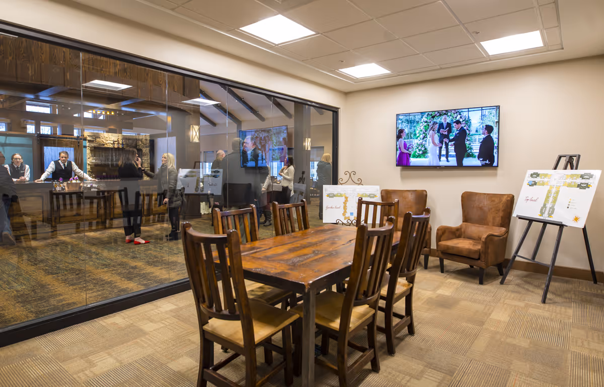 A meeting or common area in a senior living facility featuring a wooden table with six chairs, two brown armchairs, and two easels displaying floor plans. A large flat-screen TV on the wall shows a wedding scene. Through a glass wall, several people are visible in an adjacent room with a stone fireplace and wooden beams on the ceiling.