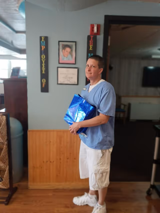An individual in blue scrubs holding a shiny blue gift bag stands in a senior living facility common area with employee recognition plaques on the wall.