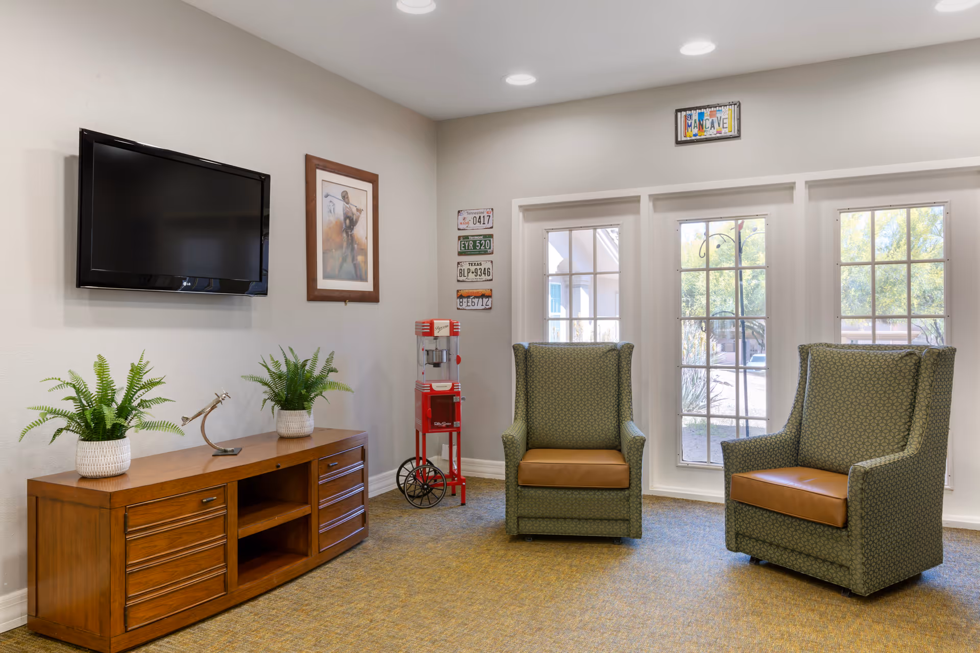 A cozy sitting area with two green upholstered armchairs with brown cushions facing a wooden TV stand with two potted plants and a decorative item. A flat-screen TV is mounted on the wall above the stand. The room has large windows letting in natural light, a framed picture on the wall, a small red popcorn machine, and a sign above the windows that reads 'MANCAVE'.