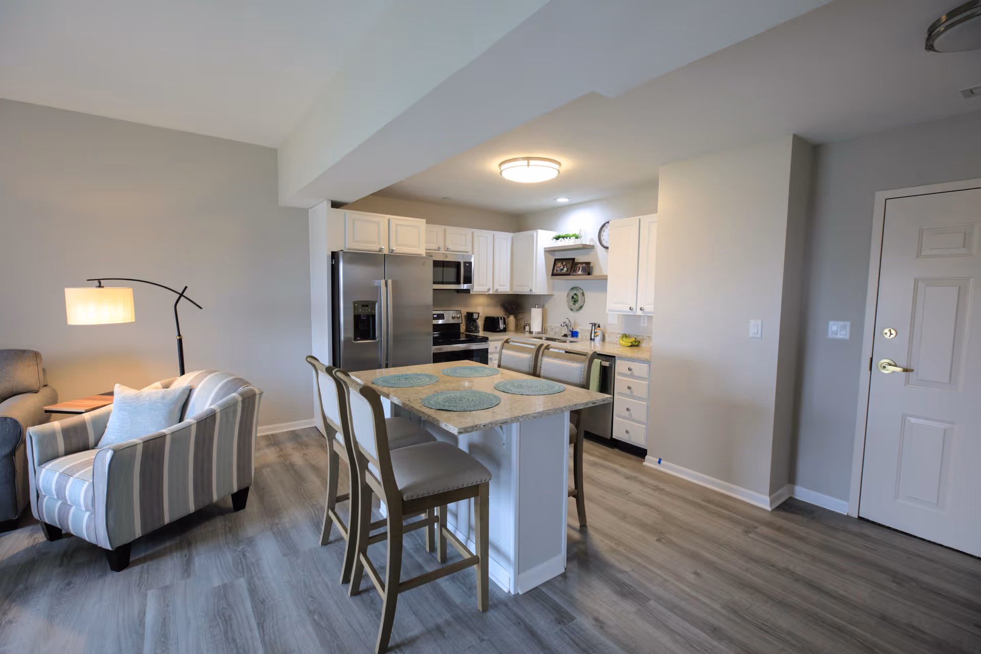 A modern kitchen and living area in a senior living facility with light wood flooring, a kitchen island with four chairs and placemats, stainless steel refrigerator and appliances, white cabinets, and a cozy seating area with a striped armchair and floor lamp.