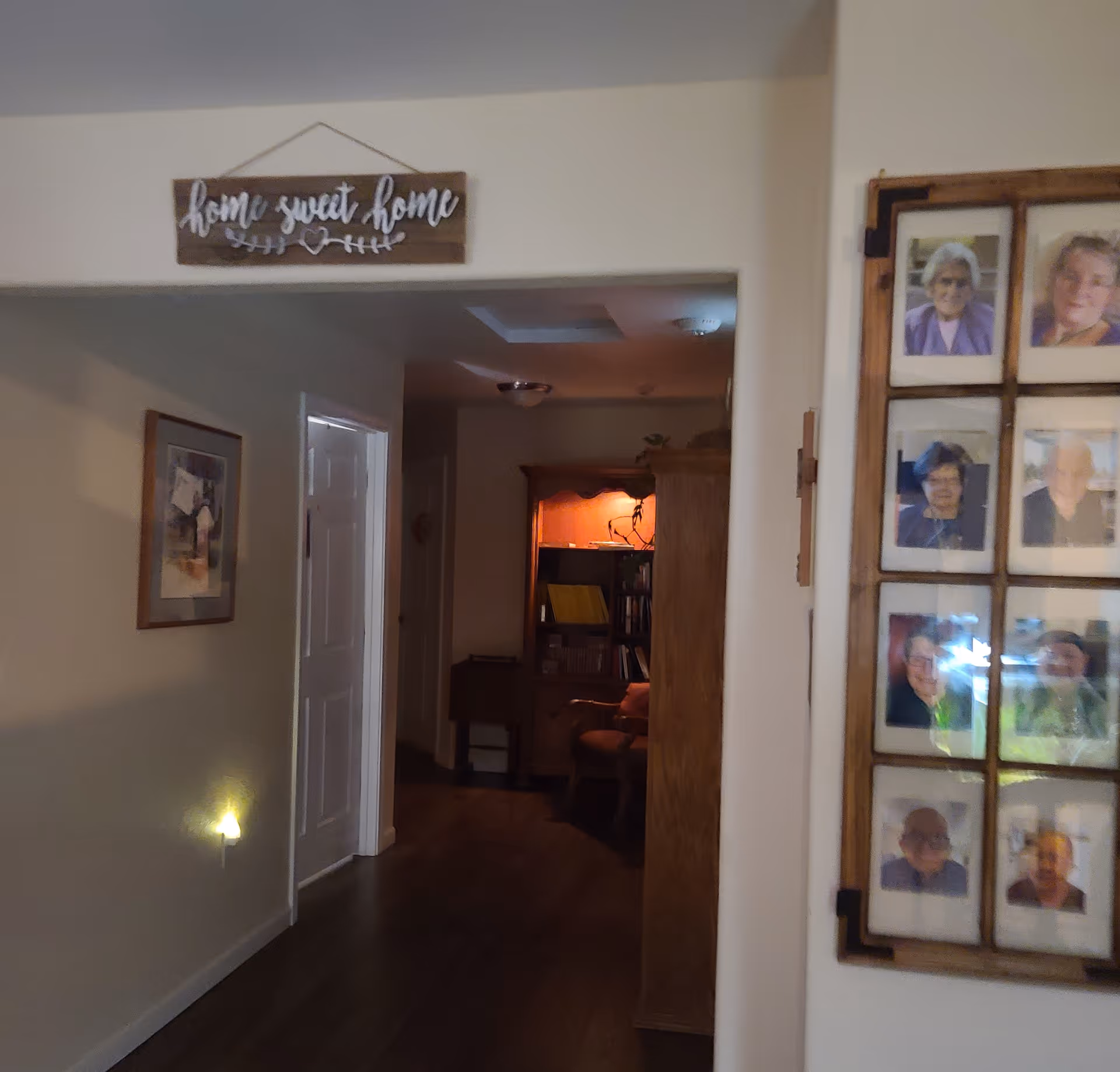 Interior hallway of a senior living facility with wooden flooring and beige walls. A wooden sign above the hallway entrance reads 'home sweet home.' On the right wall, there is a framed collage of elderly residents' photos. A softly lit room with a wooden cabinet and chair is visible at the end of the hallway.