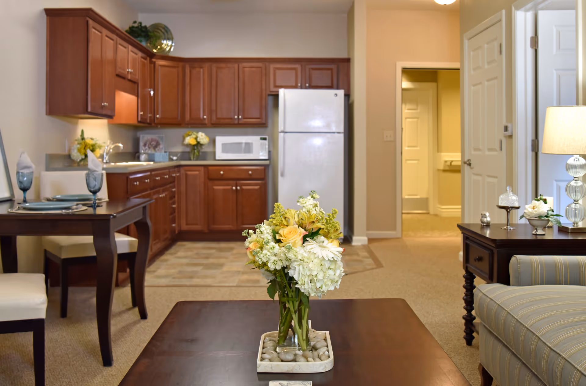 Interior view of a living space in Irene Woods Assisted Living featuring a kitchen with wooden cabinets, a white refrigerator, and a microwave. In the foreground, there is a wooden coffee table with a vase of flowers, a dining table set with plates and glasses, and a striped armchair next to a side table with a lamp and decorative items. The space is warmly lit and has beige walls and carpet.