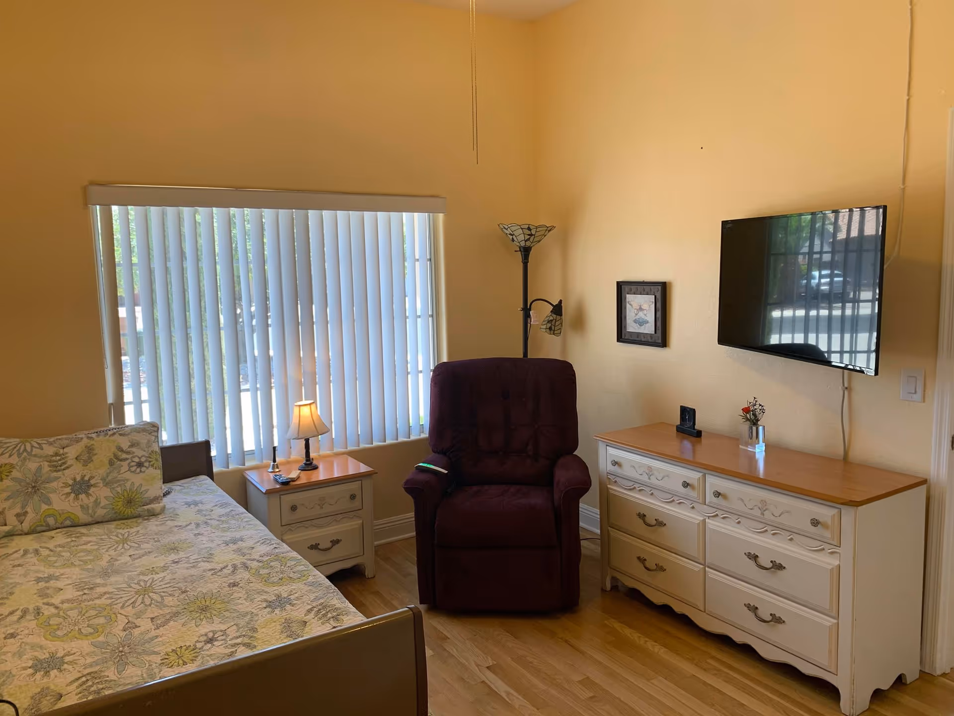 Bedroom with a bed and patterned bedding, a nightstand with lamp, a burgundy recliner, a dresser with a wall-mounted TV, and vertical blinds on the window.