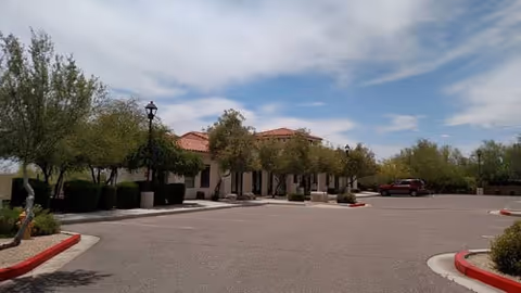 Single-story assisted living building with red tile roof, trees, and an empty parking area under a partly cloudy sky.