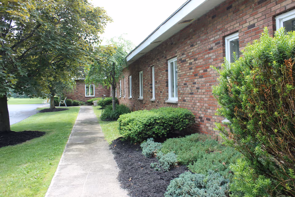 Side view of a brick building with several windows, a concrete walkway, and well-maintained landscaping including bushes, trees, and grass.