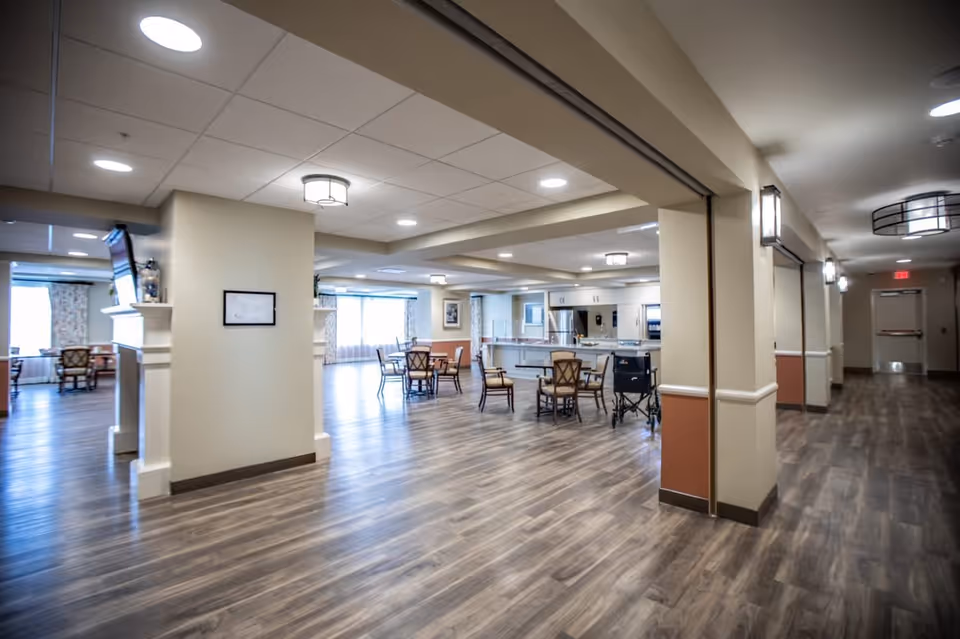 Spacious open common area with dining tables, a kitchen counter, and wood-look flooring in a senior living facility.