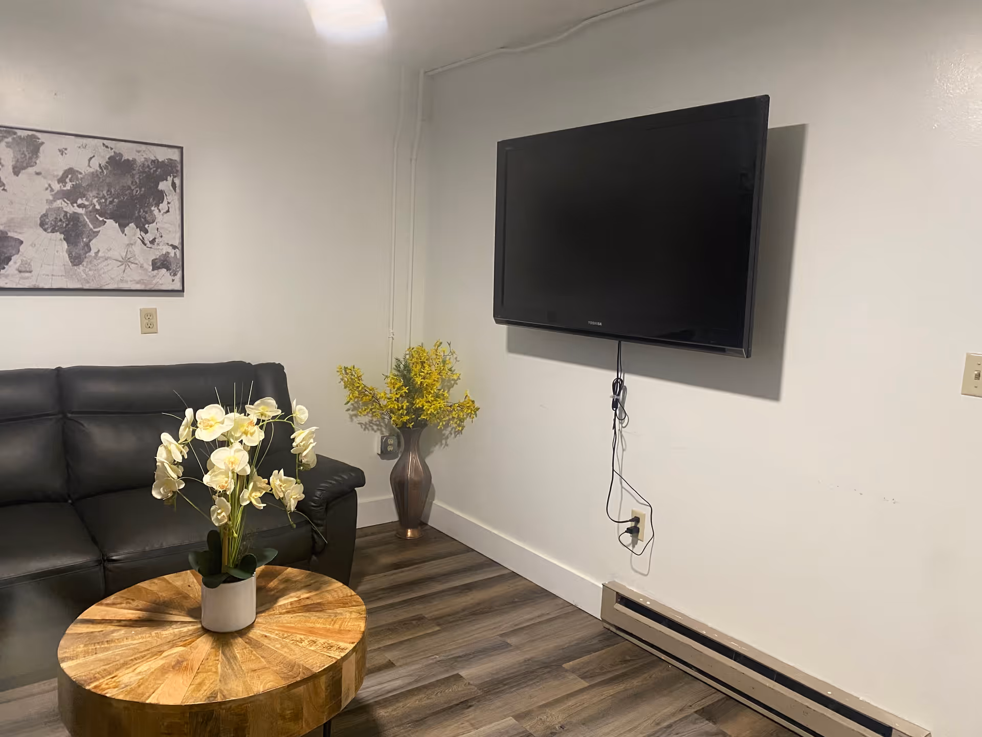 A cozy living room with a black leather couch, a round wooden coffee table with a white orchid plant on it, a wall-mounted flat-screen TV, a vase with yellow flowers in the corner, and a framed world map on the wall.