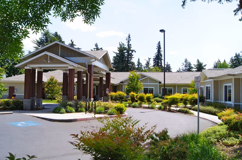 Exterior view of Avalon Care Center Portland showing a single-story building with a covered entrance supported by columns. The area is surrounded by well-maintained landscaping with bushes, trees, and a paved driveway. A 'No Parking Fire Lane' sign is visible near the entrance.