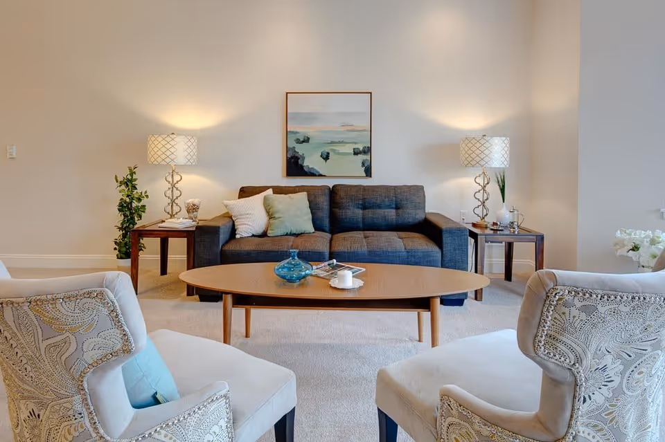 A cozy living room with a dark gray sofa adorned with light-colored pillows, two patterned armchairs facing the sofa, a wooden coffee table with decorative items, two side tables each with a lamp, a small plant on one side table, and a framed landscape painting on the wall behind the sofa.