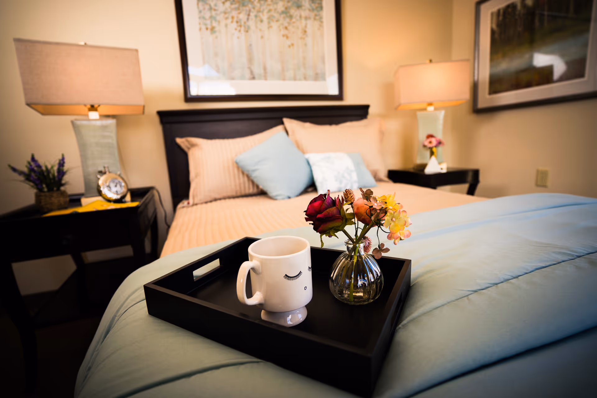 A neatly made bedroom with a black tray holding a mug and a small vase of flowers on a light blue comforter at the foot of the bed.