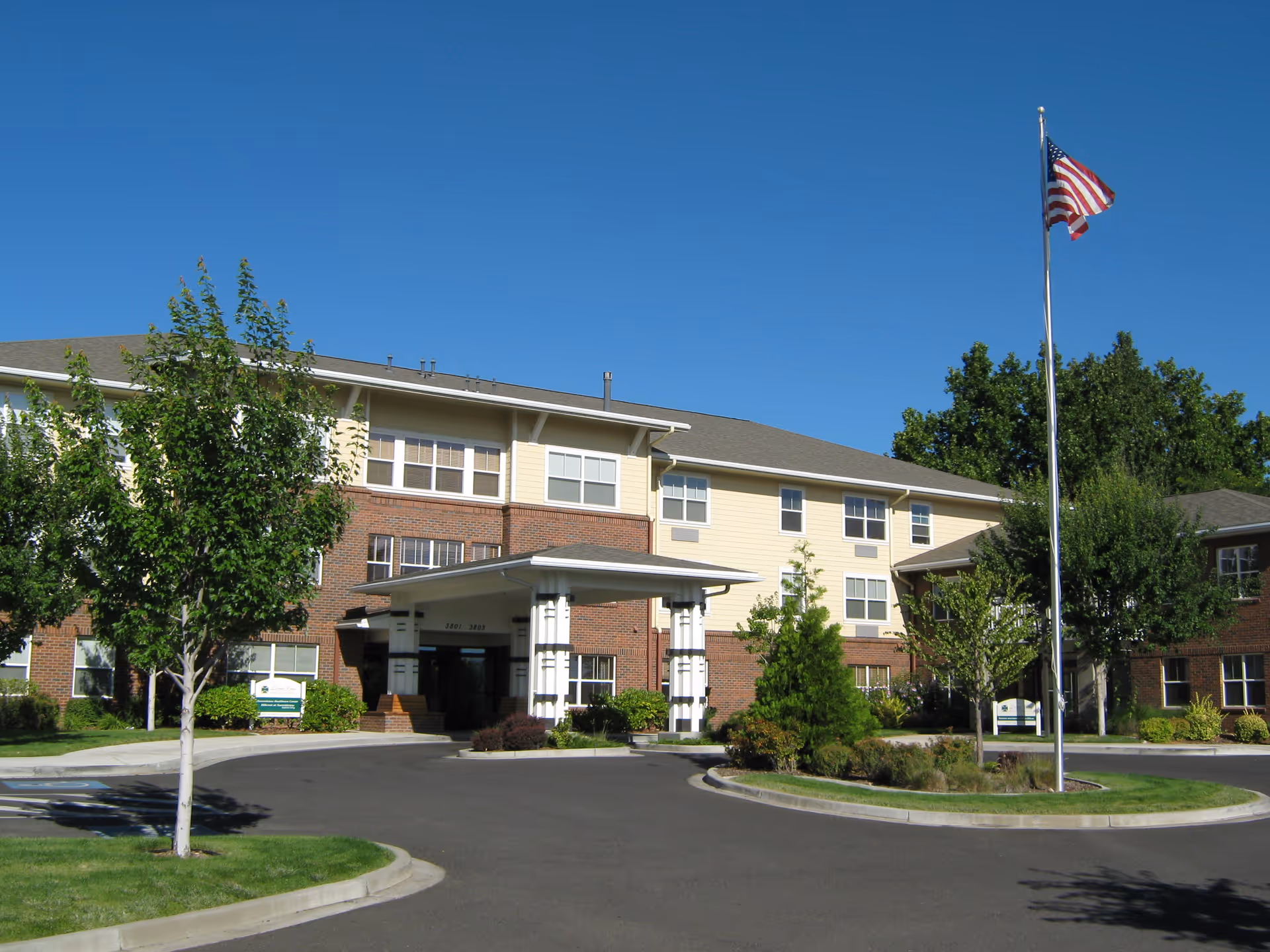 Exterior view of Hillcrest At Summitview senior living facility showing a three-story building with a covered entrance, surrounded by trees and landscaping under a clear blue sky. An American flag is flying on a flagpole near the entrance.