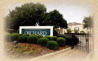 Entrance to The Orchard facility featuring a large sign with the word 'ORCHARD' surrounded by neatly trimmed bushes and trees, with a black metal gate and residential buildings visible in the background.