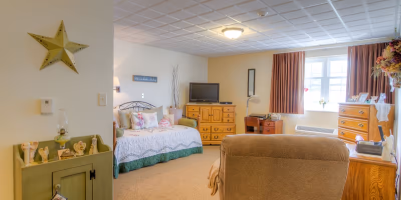 A cozy, well-lit bedroom in Carlyle Healthcare Center featuring a single bed with decorative pillows and a quilt, a wooden dresser with a TV on top, a window with brown curtains allowing natural light, a comfortable armchair, and various decorative items including a star on the wall and figurines on a small cabinet.