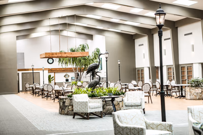 Spacious senior living common area with dining tables, lounge chairs, indoor planters and a decorative bird sculpture under skylights.