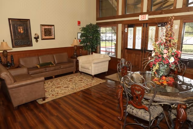 A cozy interior room with wooden flooring featuring a brown leather sofa, a matching armchair, and a beige upholstered chair around a floral area rug. There is a round glass-top dining table with ornate metal chairs and a large floral centerpiece. The room has large windows and a wooden door with glass panels, and decorative framed artwork on the walls.
