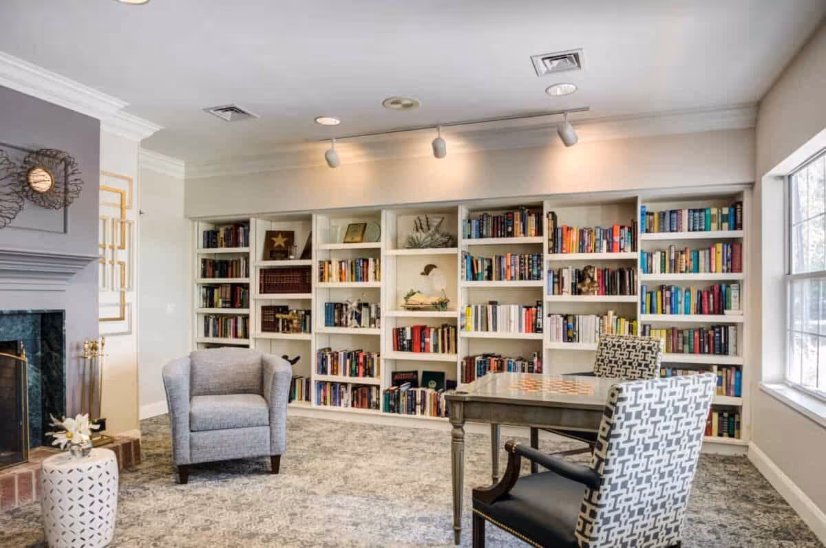 A cozy reading room with a large built-in bookshelf filled with books, a gray armchair, a table with a chessboard, and two patterned chairs. There is a fireplace on the left side and a large window on the right letting in natural light.