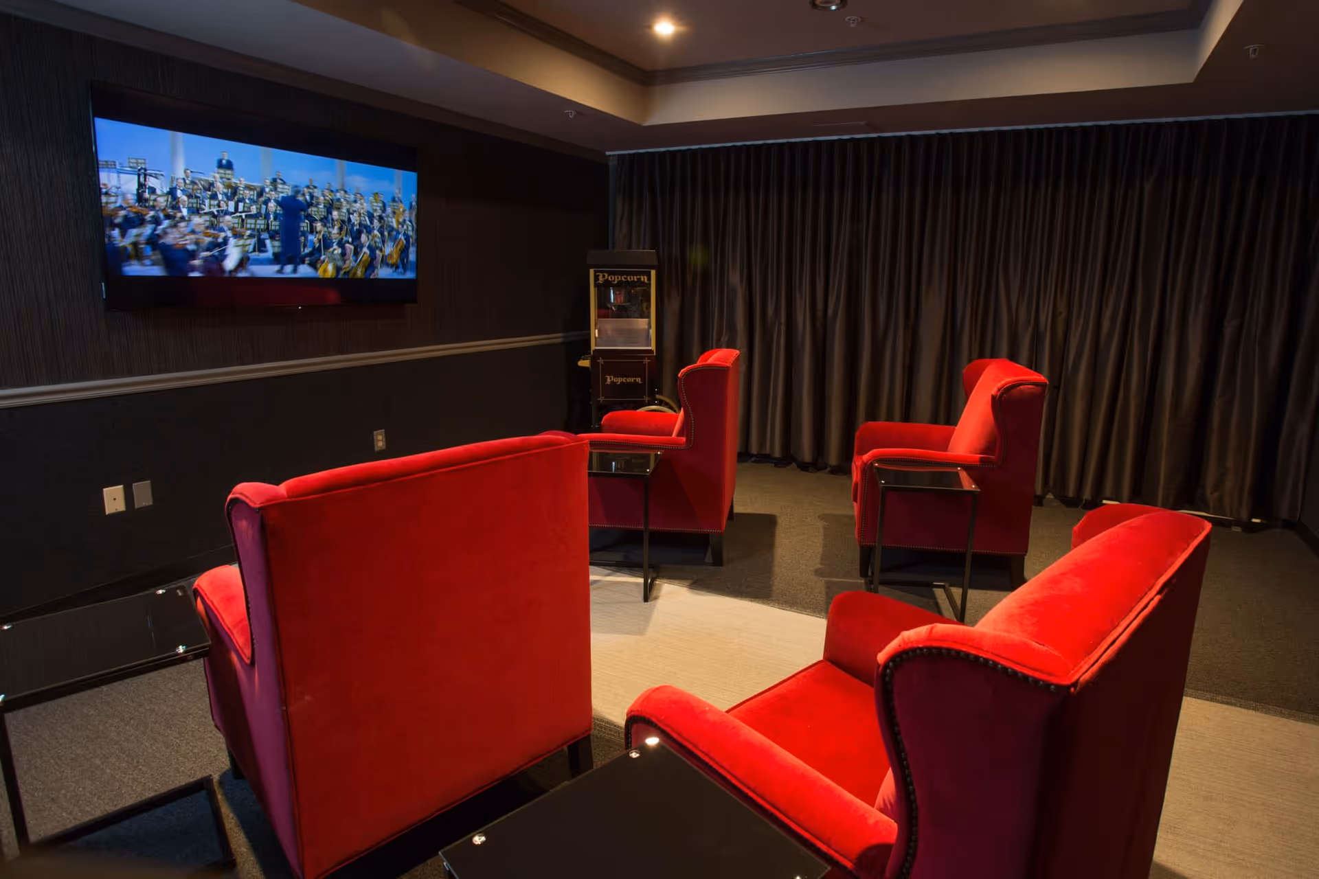 A cozy media room with four red upholstered armchairs arranged around a small table, facing a wall-mounted flat-screen TV showing an orchestra performance. There is a popcorn machine in the corner and dark curtains covering the windows.