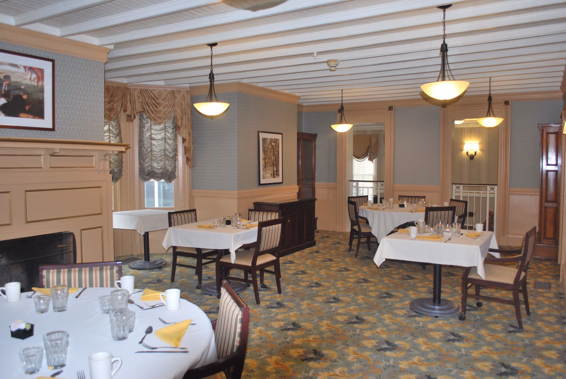 A dining room with several tables covered in white tablecloths, each set with glasses, cups, silverware, and yellow napkins. The room features patterned carpet, beige walls with wainscoting, a fireplace, framed artwork, and hanging pendant lights. Windows have decorative curtains.