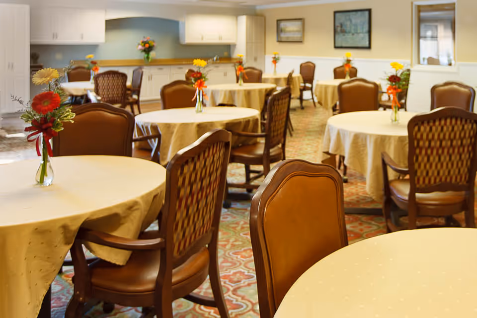 A dining room with multiple round tables covered with beige tablecloths, each adorned with a small vase of colorful flowers. The room has wooden chairs with cushioned seats and backs, patterned carpet flooring, and light-colored walls with framed artwork. Cabinets and a countertop are visible in the background.