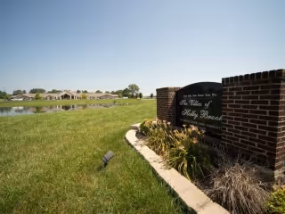 Brick entrance sign for The Villas of Holly Brook beside a grassy lawn and pond with residential buildings in the background under a blue sky.