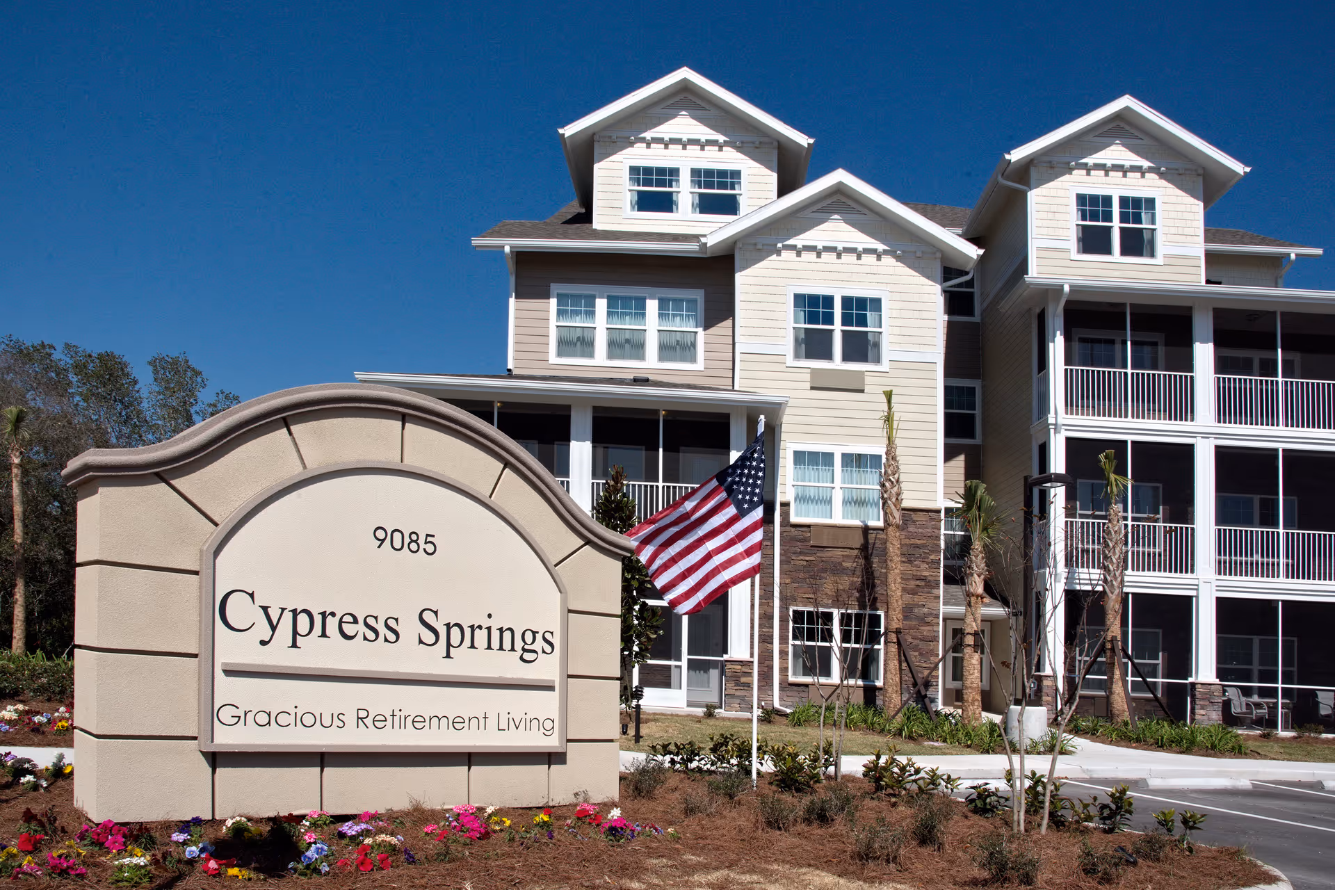 Exterior view of Cypress Springs Gracious Retirement Living building with a large sign in front displaying the facility name and address 9085. The building has multiple floors with balconies and windows, surrounded by landscaping and an American flag near the sign.