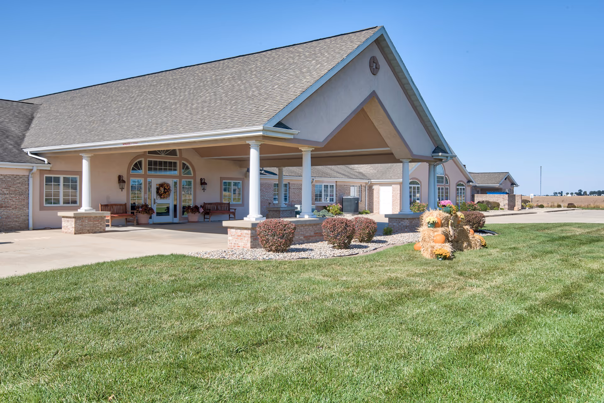 Exterior view of The Villas at St. James facility showing the front entrance with a covered driveway supported by white columns, benches near the entrance, neatly trimmed bushes, a green lawn, and a decorative fall display with pumpkins and hay bales under a clear blue sky.