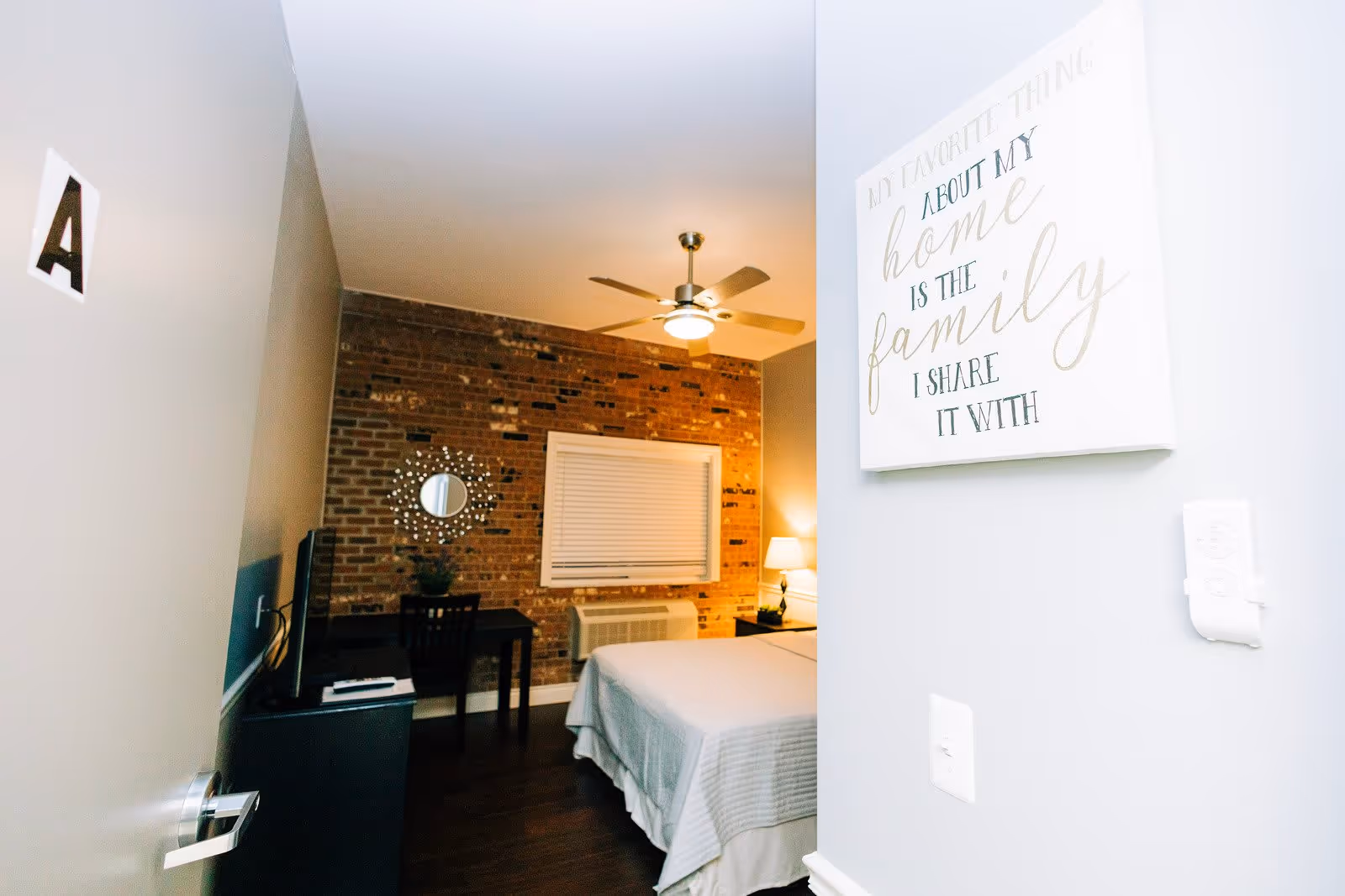 View into a bedroom with a bed covered in white linens, a ceiling fan with light, a brick accent wall, a window with closed blinds, a small desk with a chair and a decorative round mirror above it, and a TV on a stand. A wall near the entrance has a sign that reads 'My favorite thing about my home is the family I share it with.'