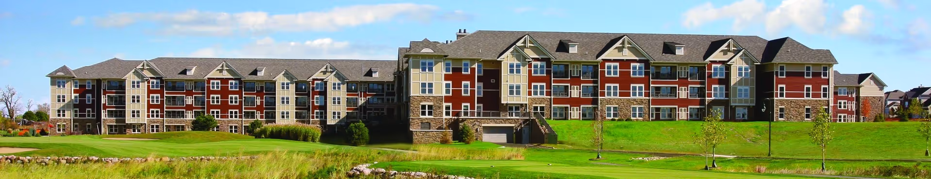 Panoramic exterior view of a long multi-story red and beige senior living building set behind a manicured green lawn.