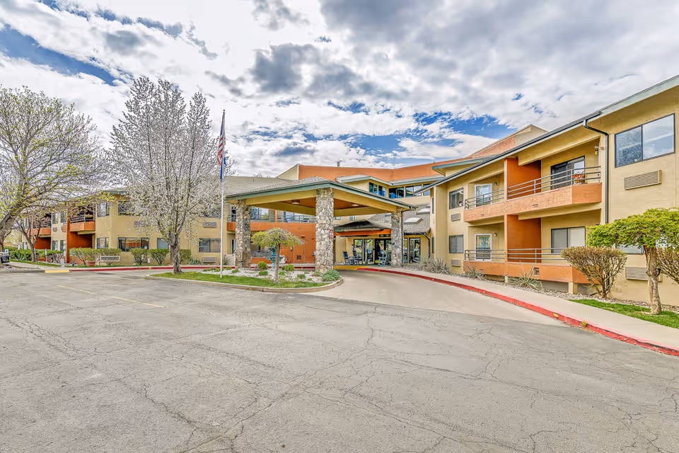 Exterior view of Solstice Senior Living at Mesa View, showing a two-story building with balconies, a covered entrance supported by stone pillars, landscaped greenery, and an American flag on a flagpole under a partly cloudy sky.