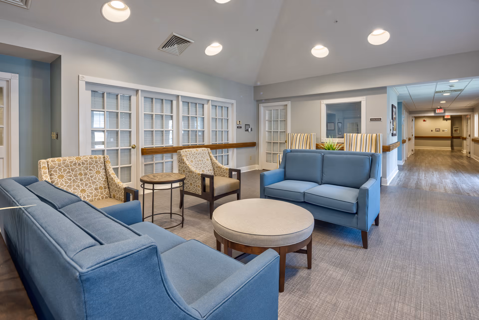 A cozy seating area in a senior living facility with two blue sofas, two patterned armchairs, a round ottoman, and a small round side table. The room has light gray walls, recessed ceiling lights, and a hallway leading to other rooms. There are white-framed glass doors and windows along one wall.