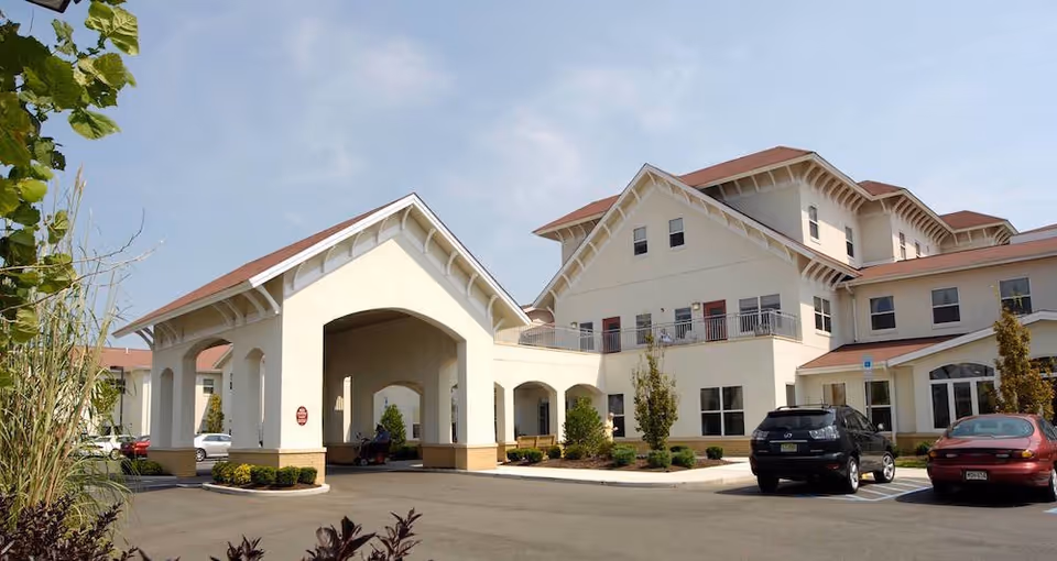 Exterior view of a senior living facility building with cream-colored walls and red roofs, featuring a covered entrance area and several parked cars in front under a clear sky.