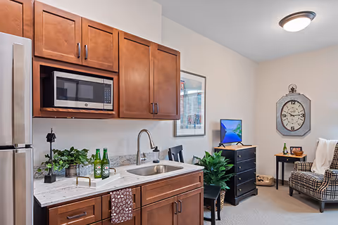 Interior view of a senior living facility kitchen and living area with wooden cabinets, a microwave, a sink, a small dining table with chairs, a potted plant, a chest of drawers with a TV on top, a large wall clock, and a plaid armchair with a white throw blanket.