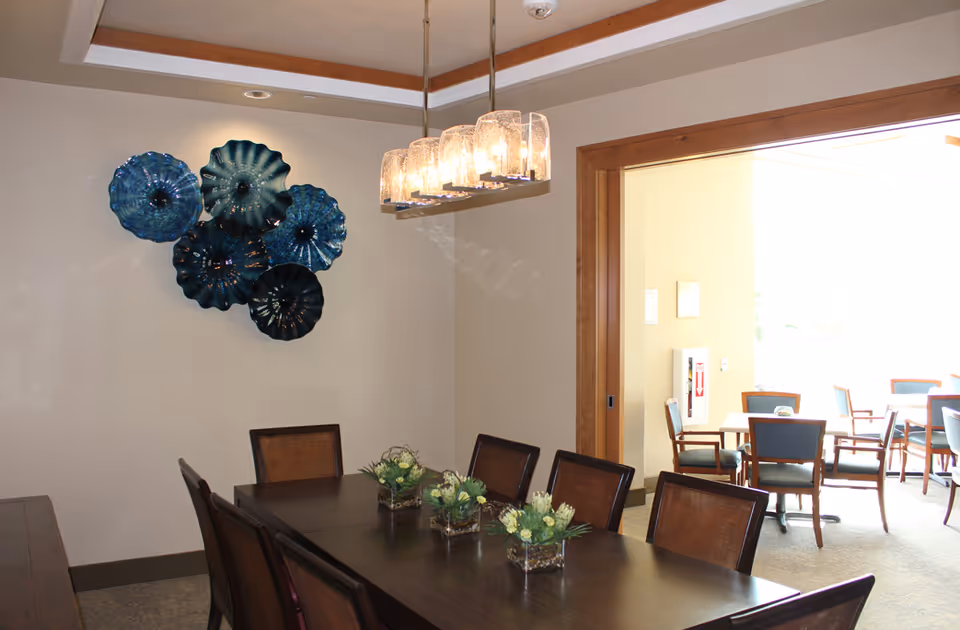 A dining room with a dark wooden table surrounded by eight chairs. Three small floral centerpieces are placed on the table. On the wall, there is a decorative arrangement of blue glass art pieces. A modern chandelier with multiple glass lights hangs above the table. In the background, there is an adjoining room with additional tables and chairs, illuminated by natural light.