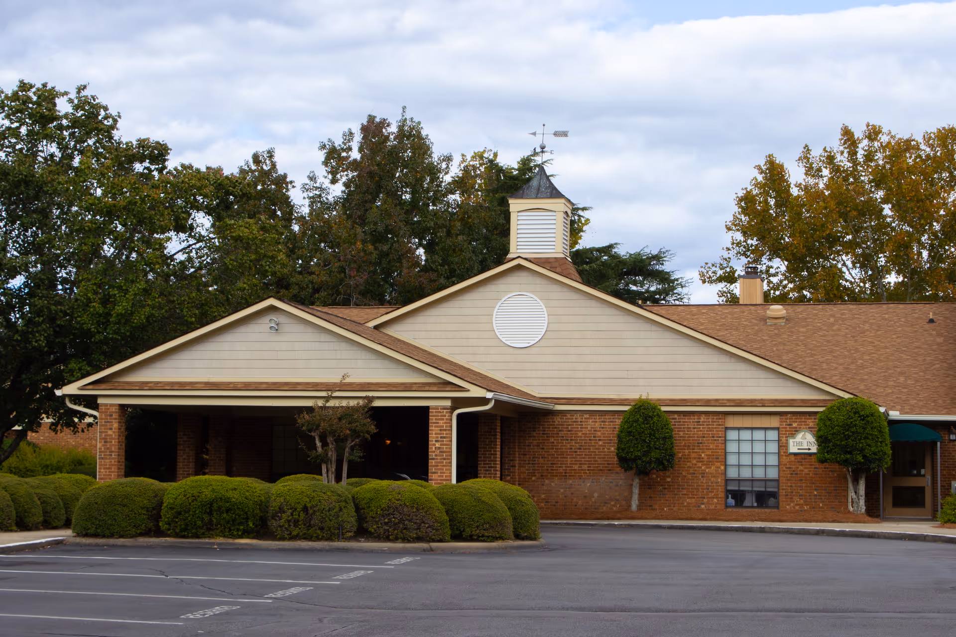 Brick single-story retirement facility front with a covered entrance, manicured shrubs, and trees under a cloudy sky.