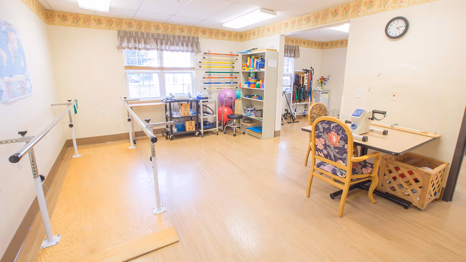 Bright therapy/exercise room with parallel walking bars, equipment shelves, and a table with chairs.