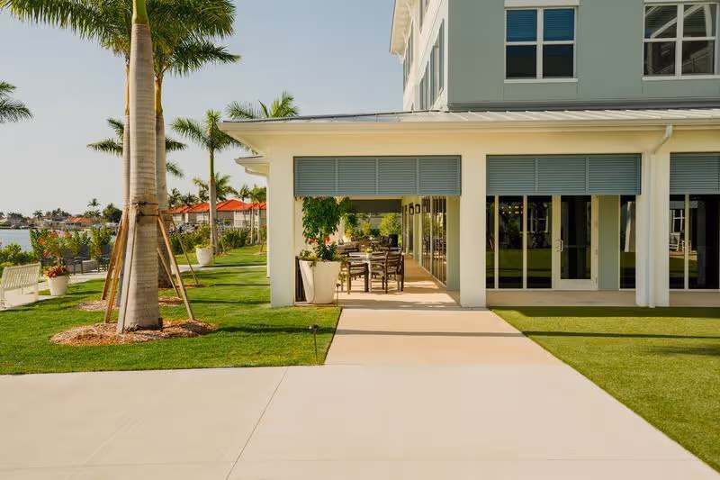 Outdoor patio and walkway beside a modern coastal building with palm trees and waterfront views.