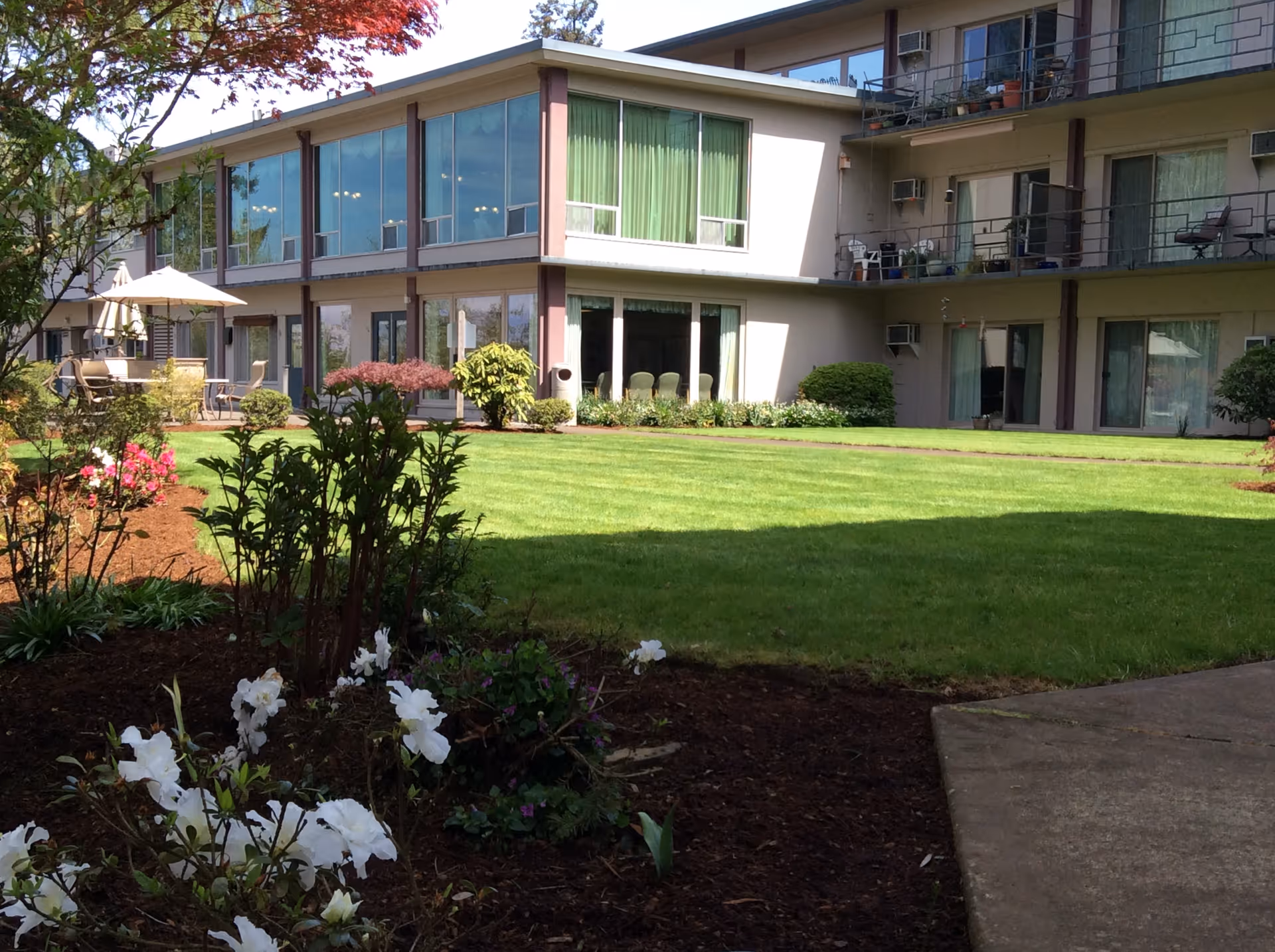 Outdoor view of a multi-story retirement facility building with large windows and balconies. The foreground features a well-maintained lawn, flower beds with white and pink flowers, shrubs, and a patio area with tables and umbrellas. Trees and clear blue sky are visible in the background.