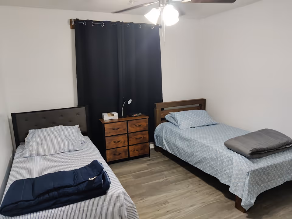 A bedroom with two single beds, each made with patterned bedding and pillows. Between the beds is a wooden dresser with six drawers, a small lamp, and a tissue box on top. The room has light-colored walls, wood flooring, a ceiling fan with lights, and a window covered by dark curtains.