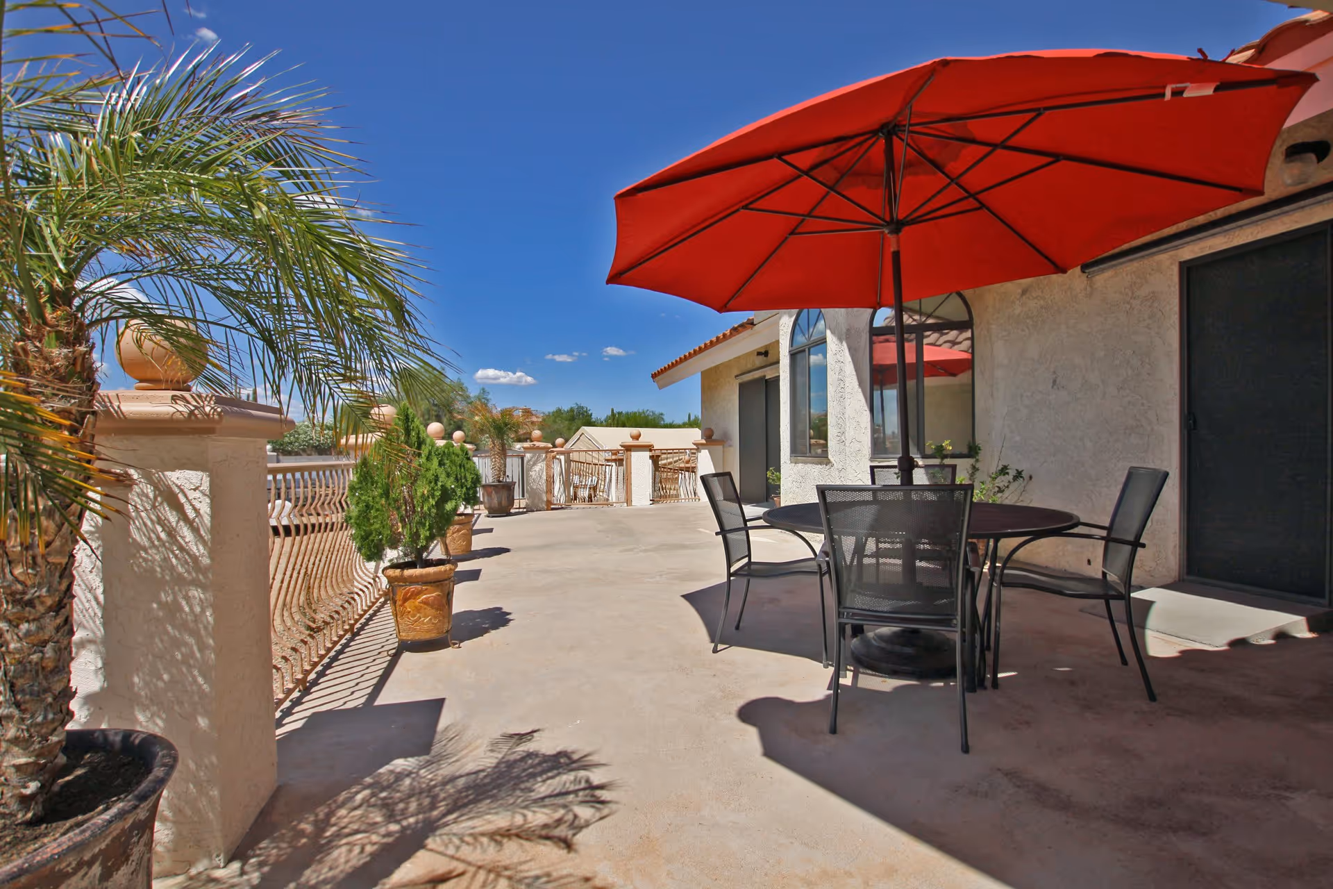 Outdoor patio area with a round table and four black metal chairs under a large red umbrella. The patio is surrounded by a decorative railing and several potted plants, including palm trees and small shrubs. The sky is clear and blue.