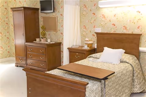 A senior living facility bedroom with a wooden bed featuring a beige patterned bedspread and a white pillow. There is a wooden nightstand with a lamp and some items on it next to the bed. Across from the bed is a wooden dresser with drawers and a cabinet, with a small TV mounted on the wall above it. The walls have floral wallpaper, and the floor is light-colored.