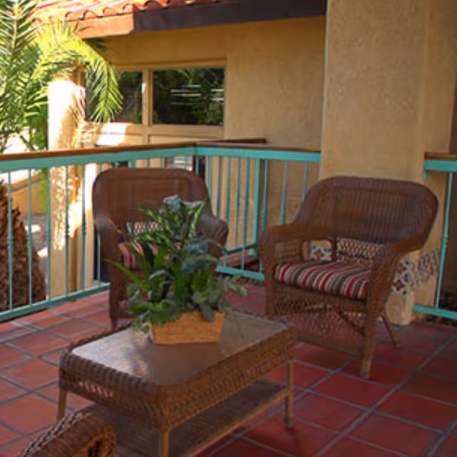 Outdoor patio area with two wicker chairs featuring striped cushions and a wicker table with a potted plant on top. The patio has terracotta tile flooring and a turquoise railing, with a palm tree visible in the background.